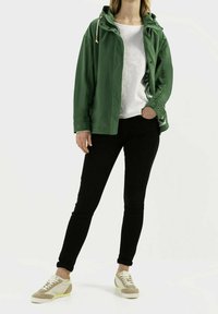 Personne portant une veste verte par-dessus une chemise blanche avec un pantalon noir et des baskets beiges, debout avec une main dans la poche.