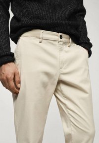 Beige Baumwollhosen mit flachem Bund, seitlichen Taschen und einem schwarzen Knopfverschluss, kombiniert mit einem strukturierten schwarzen Strickpullover.