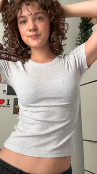 Light grey ribbed cotton t-shirt with short sleeves, fitted design, round neckline, worn with black pants. Background features posters and plants.