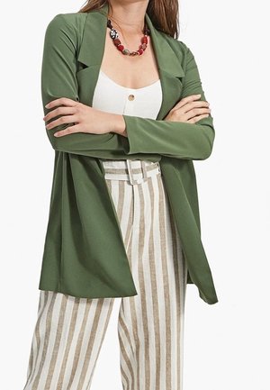 Woman with crossed arms wearing an olive green blazer, white buttoned tank top, striped beige and white high-waisted pants, and a red beaded necklace.