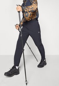 Pantalons de randonnée bleu foncé en tissu durable, dotés de plusieurs poches, associés à des chaussures de randonnée noires. Des bâtons de trekking réglables sont tenus.