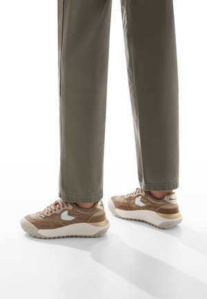 Sneakers beige con lacci, caratterizzati da una parte superiore testurizzata, un accento del logo bianco e una suola intermedia spessa bianca. Indossati con pantaloni ampi color verde oliva.