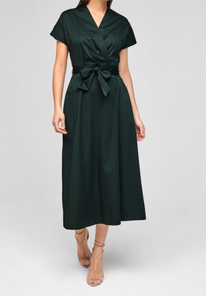 Frau trägt ein dunkelgrünes Midikleid mit kurzen Ärmeln, V-Ausschnitt, Taillengürtel und beigen High Heel Sandalen mit Knöchelriemen, steht vor einem grauen Hintergrund.