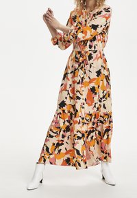 Een lange, vloeiende jurk met een crème basis en abstracte patronen in roze, oranje en zwart. Heeft een strakke taille en een gelaagde rok, gecombineerd met witte laarzen.