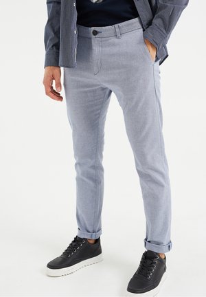 Homme portant un pantalon slim gris clair avec les revers retroussés, une chemise bleu marine, une veste à carreaux et des baskets noires avec des semelles blanches.