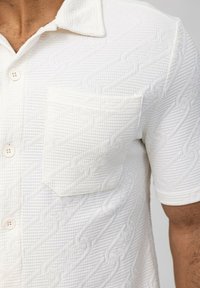 Chemise blanche à manches courtes en tissu texturé avec un motif matelassé prononcé, dotée d'une poche poitrine et de boutons ronds.