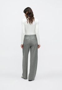 Femeie cu păr lung și închis la culoare, purtând un top alb cu mâneci lungi și pantaloni largi cu dungi gri, stând în picioare pe un fundal simplu.