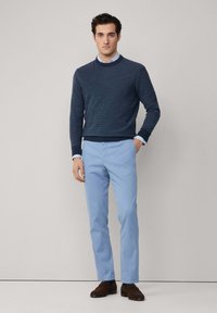 Mornarsko plavi pulover teksture preko svijetloplavog košulje s ovratnikom, uparen sa svijetloplavim hlačama i smeškim suedinim cipelama.