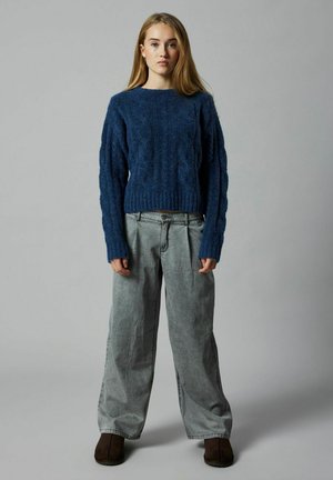 Maglione blu lavorato a maglia con motivo a treccia, con maniche lunghe e colletto tondo. Abbinato a pantaloni grigi larghi e scarpe marroni scure.