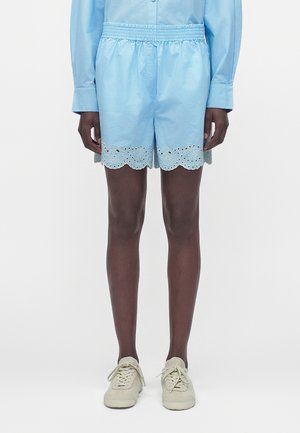 Person wearing light blue elastic waist shorts with eyelet lace trim and beige sneakers, standing against a plain white background.