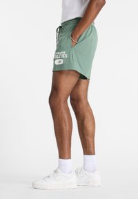 Grønne mesh treningsshorts med sidelommer, snøring i livet, og logoen "New Balance Athletics". Kombinert med hvite joggesko og sokker.
