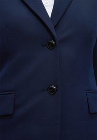 Blazer bleu marine avec deux boutons noirs, col cranté et poches à rabat sur une chemise blanche.