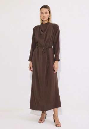 Γυναίκα φοράει καφέ φόρεμα μακρυμάνικο μέχρι τον αστράγαλο με δεμένη ζώνη στη μέση και μπεζ ανοιχτά πέδιλα, ποζάροντας μπροστά σε απλό λευκό φόντο.