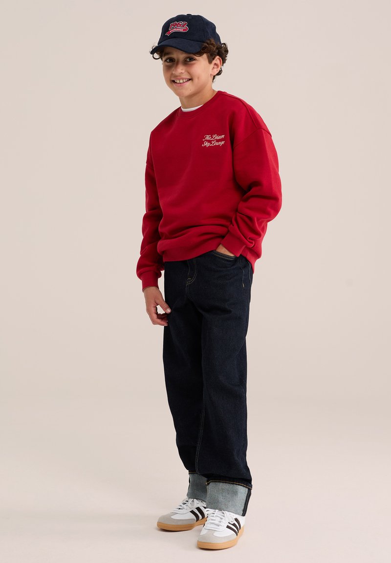 Jeune garçon souriant portant un sweat-shirt rouge, un jean foncé retroussé, des baskets blanches et une casquette bleu marine avec un logo rouge.