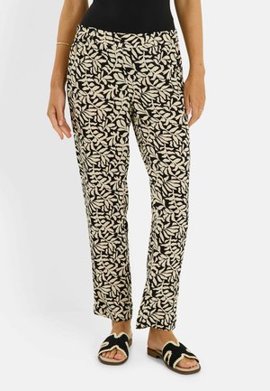 Femme portant un pantalon à motif feuilles noir et beige avec des sandales tissées noir et beige à bouts ouverts sur un fond uni.