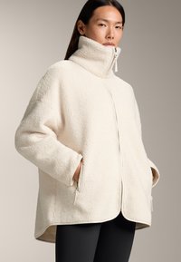 Giacca in pile crema con colletto alto, zip completa e tasche laterali. Presenta una texture morbida e orlo arrotondato per un fit rilassato.