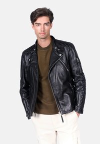 Czarna skórzana kurtka motocyklowa z pikowanymi ramionami, asymetrycznym zamkiem i srebrnymi okuciami. Nosi się ją na teksturowanej oliwkowej koszuli.