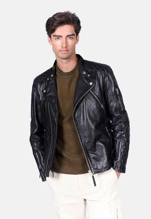 Chaqueta de motocicleta de cuero negro con hombros acolchados, cremallera asimétrica y herrajes plateados. Lleva puesta sobre una camisa texturada de color verde oliva.