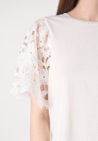 Biała koszulka z kwiatowymi koronkowymi rękawami o misternych zdobieniach. Materiał jest gładki, a koronka ma falowany brzeg.