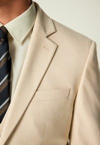 Blazer beige con corte entallado, solapas de muesca y un bolsillo en el pecho. Debajo, se ve una camisa de textura verde claro y una corbata azul marino con estampado.