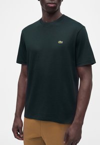 Modèle masculin portant un t-shirt noir à manches courtes avec un petit logo crocodile vert sur la poitrine, associé à un pantalon beige.