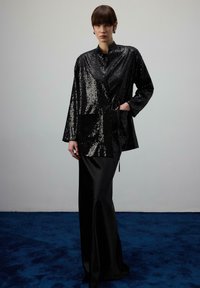 Žena s krátkymi hnedými vlasmi, oblečená v lesklej čiernej sequinovej bunde cez dlhú čiernu saténovú sukňu, stojaca na modrej kobercom pokrytej podlahe.