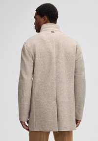 Beige wolmix jas met een visgraatpatroon, hoge kraag en een rechte snit. Model wordt van achteren getoond met ontspannen handen.