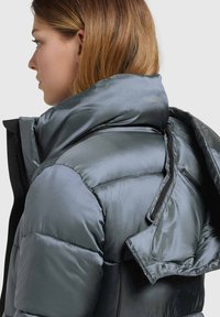 Chaqueta acolchada gris de plumón con cuello alto y abertura con cremallera, que presenta una textura lisa y brillante, además de detalles de hardware ocultos.