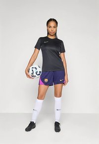 Camiseta preta de manga curta da Nike combinada com calções do FC Barcelona; acentos azuis e rosas. Meias brancas até ao joelho, sapatos de futebol pretos, segurando uma bola de futebol.