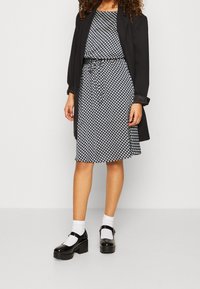 Veste noire portée sur une robe noire et blanche à motifs avec une taille cintrée, associée à des chaussures Mary Jane noires brillantes et des chaussettes blanches.