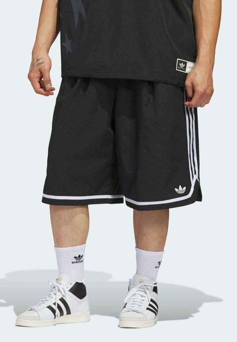 Shorts de sport noirs avec des rayures blanches, offrant une coupe décontractée et un logo Adidas. Associés à des baskets blanches et des chaussettes.
