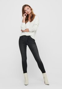 Witte gebreide trui met een v-hals, gecombineerd met hoge zwarte skinny jeans. Model draagt crèmekleurige enkellaarsjes. Soepele textuur, casual ontwerp.