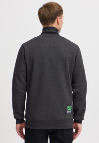 Sudadera gris con cuello alto, puños acanalados y un pequeño estampado gráfico verde en la parte posterior. Tejido texturizado, corte relajado.