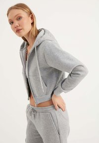 Felpa grigia con zip, cappuccio, tasche frontali e polsini a costine. Abbinata a pantaloni grigi coordinati, realizzati in morbido tessuto testurizzato.