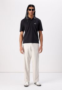 Polo negru cu mânecă scurtă și logo, asortat cu pantaloni bej deschis și adidași alb-negru. Modelul poartă ochelari de soare.