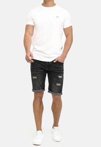Witte katoenen t-shirt met een klein logo op de borst, gecombineerd met donkere denim shorts met versleten details en opgerolde mouwen, gedragen met witte sneakers.