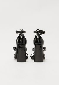 Sandales à talons en cuir verni noir avec un talon bloc rectangulaire, une sangle à la cheville et un design à bout ouvert. Texture lisse avec une finition brillante.