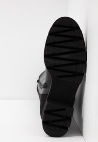 Semelle d'une botte noire avec des motifs de bande de roulement profonds et angulaires, reposant contre une surface d'angle blanche.
