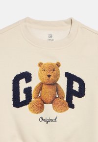 Bēšs mazuļa džemperis ar mīkstu lāča rotaļlietu starp teksturētām tumši zilām burtiem "G" un "P" un vārdu "Original" zemāk.