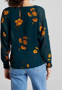 Blouse fleurie en tissu vert foncé avec des fleurs orange, coupe décontractée, dos à découpes en forme de goutte, et poignets élastiqués. Portée avec un jean bleu clair.