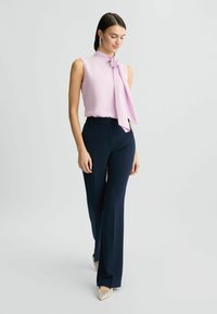Lichtroze mouwloze top met een strikdetail, gecombineerd met hoge taille marineblauwe broeken. Model draagt puntige, versierde nude hakken.