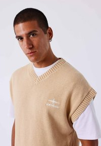 Suéter sin mangas tejido en color beige con escote en V, que presenta un cuello acanalado y un detalle de logotipo en el pecho, superpuesto a una camisa blanca.