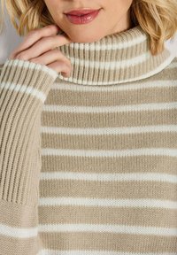 Beige gestreifter Rollkragenpullover mit breiten weißen Streifen, geripptem Material und hohem Kragen. Die Ärmel sind leicht locker sitzend.