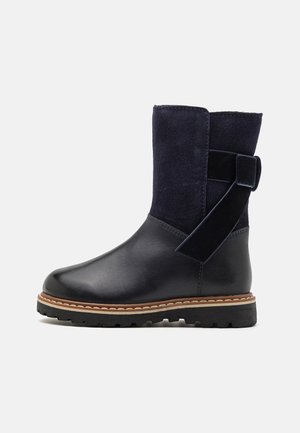 Botas de tornozelo azul marinho com bico de couro liso e parte superior em camurça texturizada, apresentando uma faixa larga e detalhe de fivela na parte de trás, sola robusta.