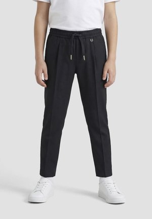 Persoon die zwarte broek met trekkoord, witte sokken, witte sneakers en een wit T-shirt met korte mouwen draagt, staand tegen een effen achtergrond.