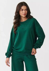 Zielony bawełniany sweatshirt z prążkowanymi mankietami i dołem, z teksturowanym logo "AKADAMIA" na przodzie. Luźny krój i miękka faktura.