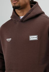 Bruine hoodie gemaakt van zachte stof met een grote voorzak, een capuchon met trekkoord en twee witte opdrukken met de teksten "STRONGER" en "chance."