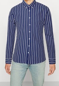 Uomo che indossa una camicia a maniche lunghe, blu navy con righe verticali bianche e bottoni, abbinata a jeans azzurri.