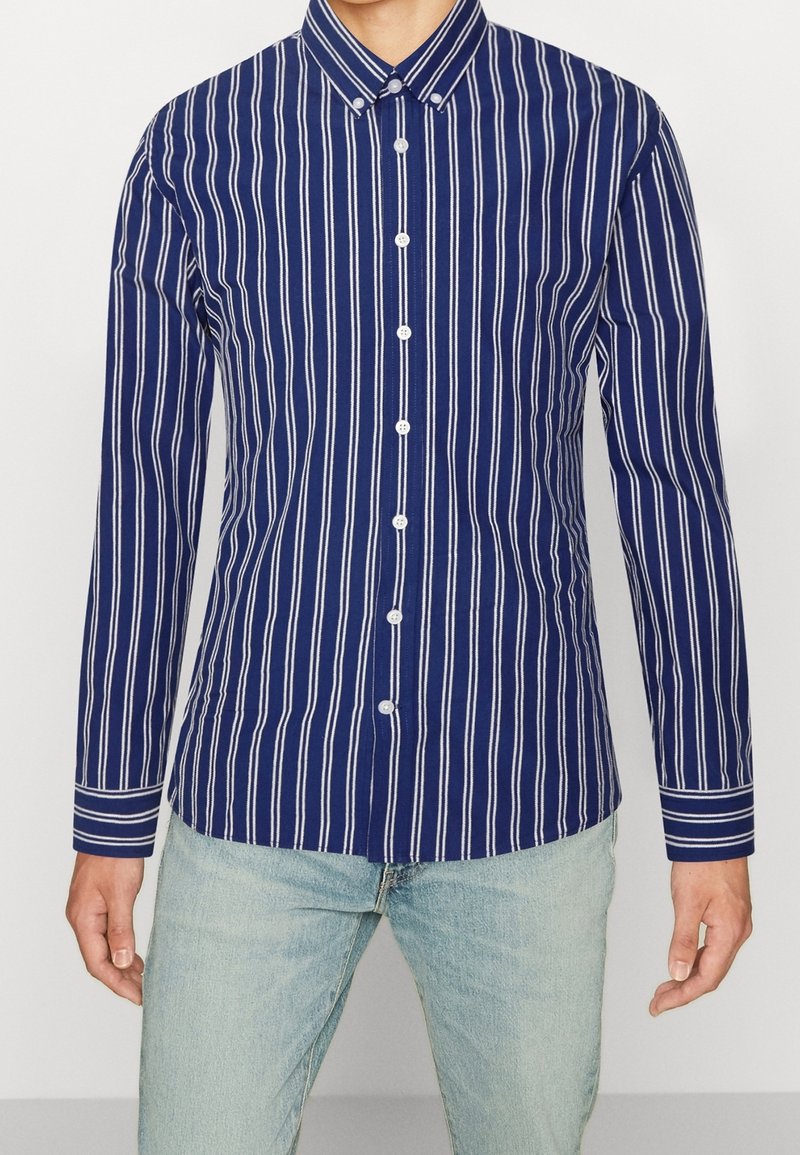 Uomo che indossa una camicia a maniche lunghe, blu navy con righe verticali bianche e bottoni, abbinata a jeans azzurri.