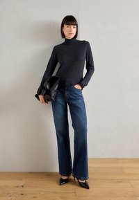 Mørk grå genser med høy krage, blå vide jeans med frynser på kanten, og svarte spisse hæler, holdt med en svart veske mot en nøytral bakgrunn.